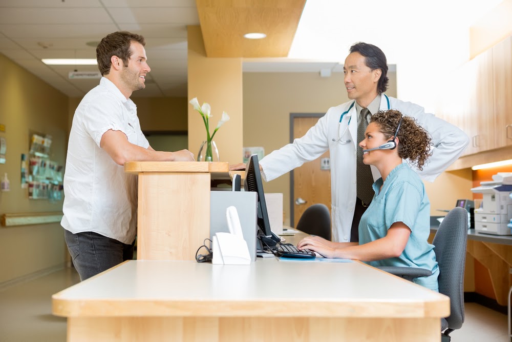 Man standing at doctor office desk talking to doctor and assistant
